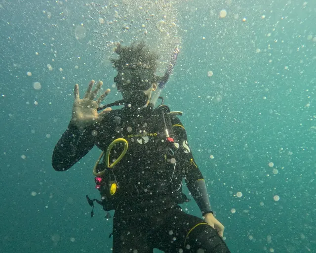 A diver in scuba gear waving underwater, surrounded by bubbles