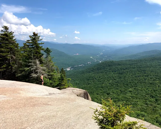A hiker on a granite ledge overlooking a forested valley in New Hampshire